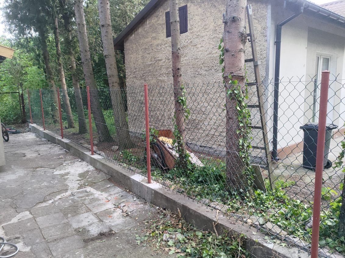 Częściowo ukończony montaż ogrodzenia z siatki z czerwonymi słupkami na betonowej podmurówce, widoczne drzewa i budynek w tle, teren po budowie.