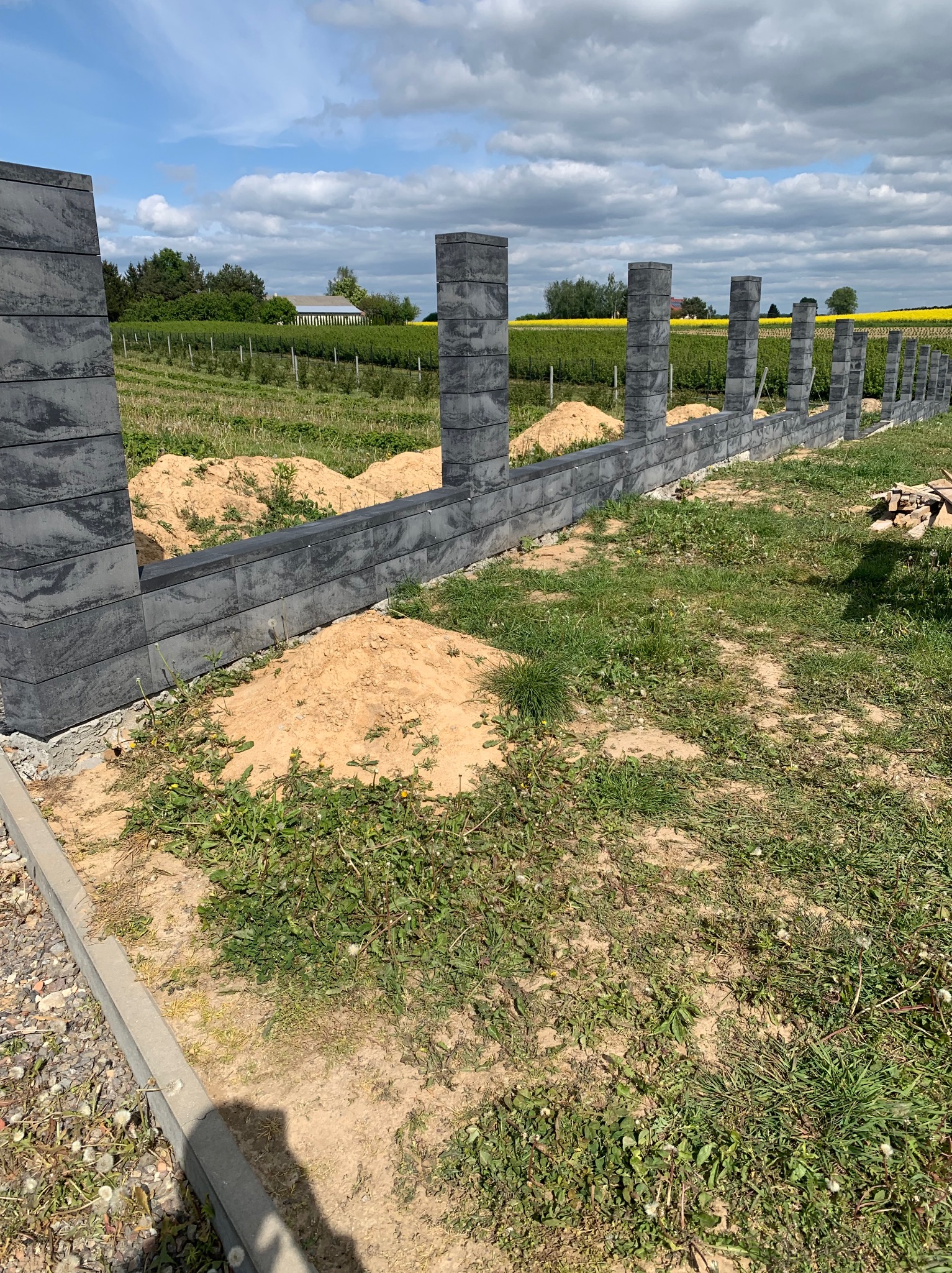 Wznoszenie ogrodzenia modułowego z szarych bloczków betonowych na tle zielonego pola i błękitnego nieba z chmurami. Widoczne słupki i fragmenty muru.