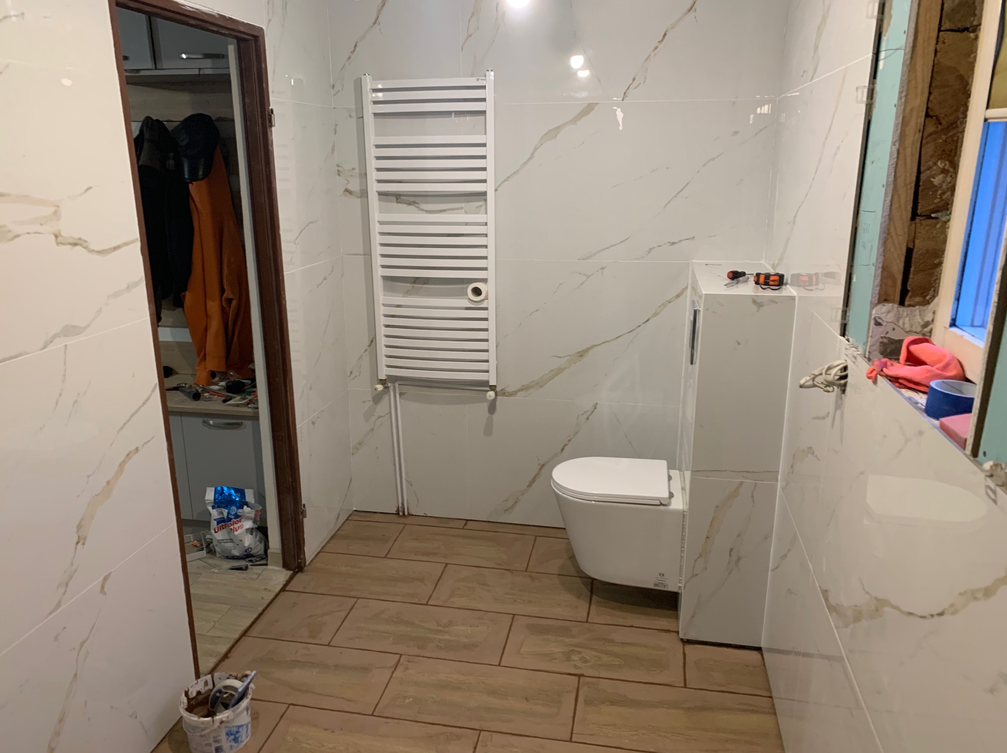 Remont łazienki: białe płytki imitujące marmur na ścianach, brązowe płytki na podłodze, podwieszana toaleta, grzejnik drabinkowy. Widoczne narzędzia i materiały budowlane.