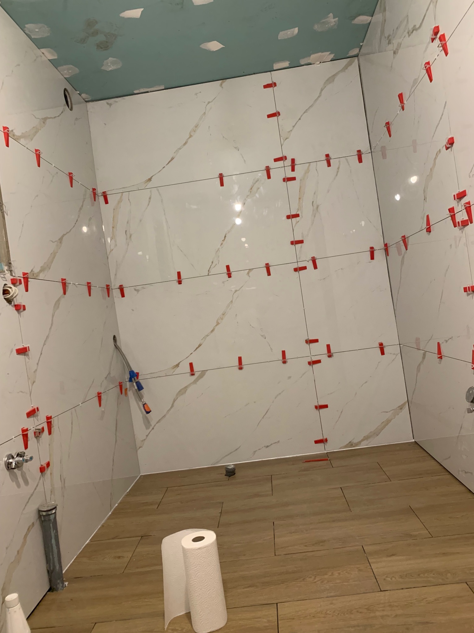 Łazienka w trakcie wykańczania: białe płytki imitujące marmur na ścianach, brązowe płytki podłogowe. Widoczne czerwone klipsy do poziomowania płytek i rolka papieru.
