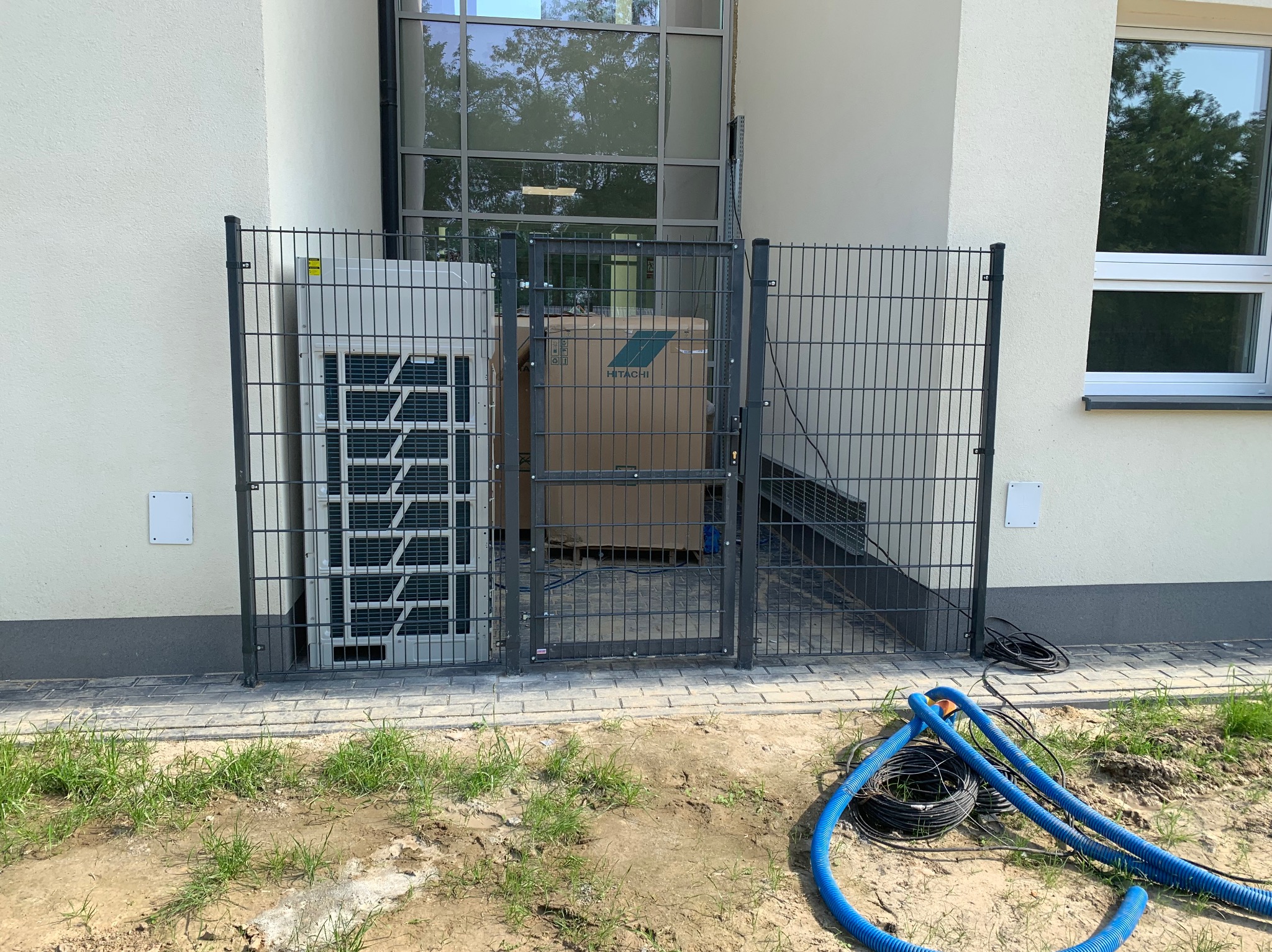 Szara brama panelowa z siatki, chroniąca klimatyzator i kartonowe pudło Hitachi. Na pierwszym planie zwój niebieskich kabli. Trawa i kostka brukowa przy budynku.