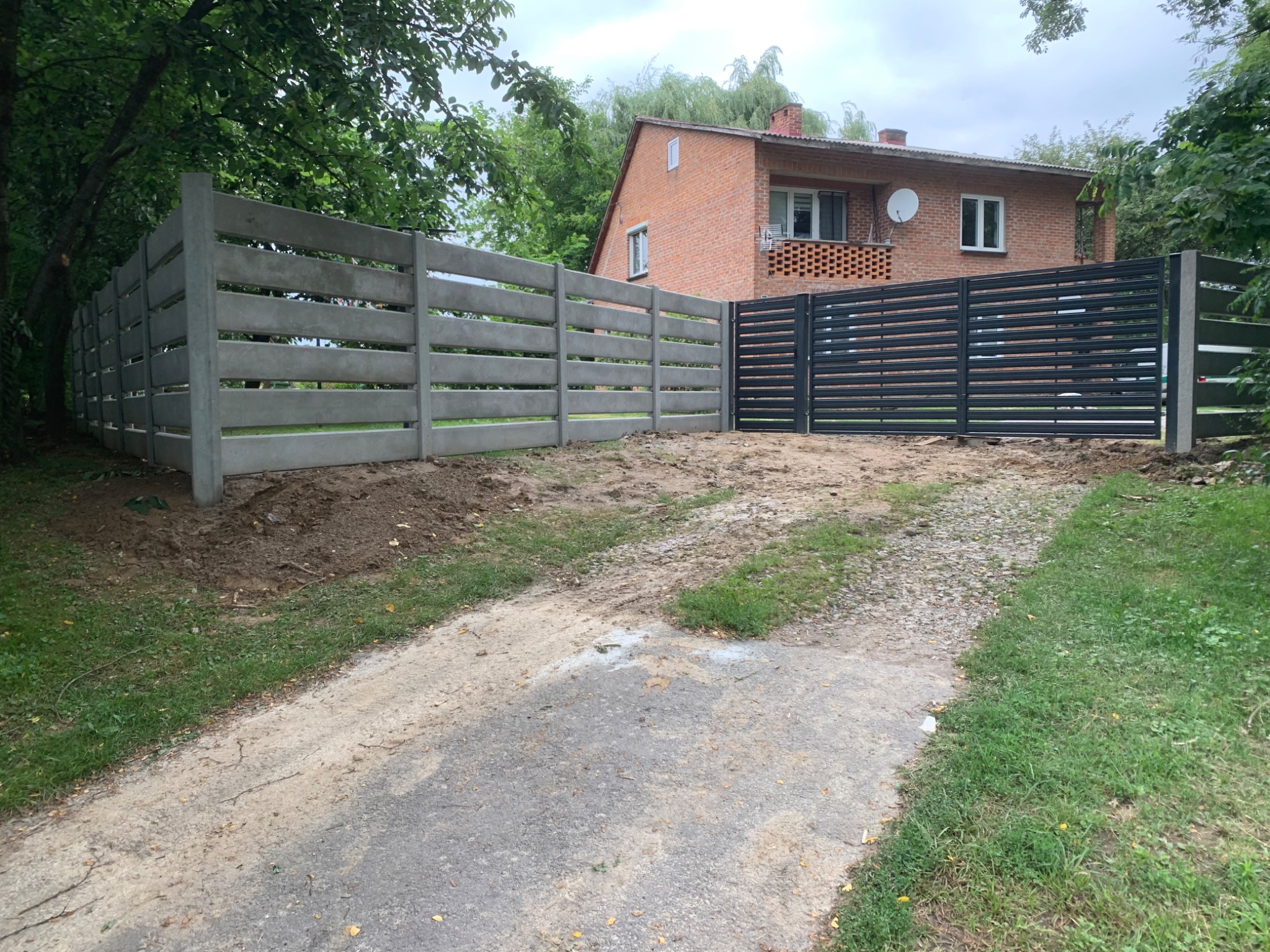 Nowoczesne ogrodzenie betonowe i brama panelowa w kolorze antracytowym przed domem z czerwonej cegły, otoczone zielenią. Elementy ogrodzenia poziome.