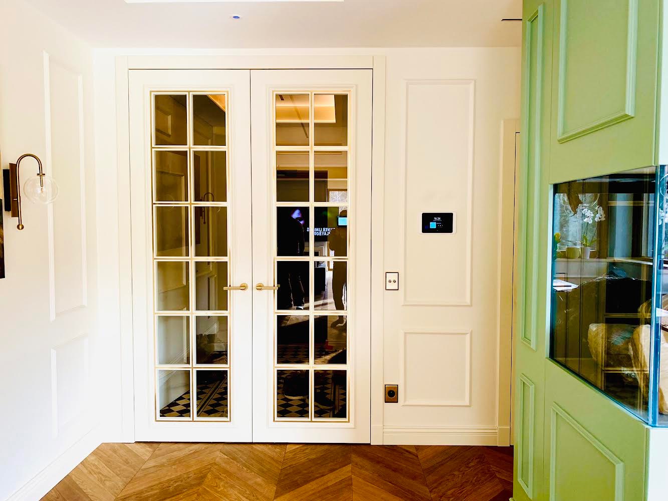 Białe, przeszklone drzwi dwuskrzydłowe z złotymi ramkami, zamontowane w klasycznym wnętrzu z drewnianą podłogą ułożoną w jodełkę i zielonymi elementami dekoracyjnymi.