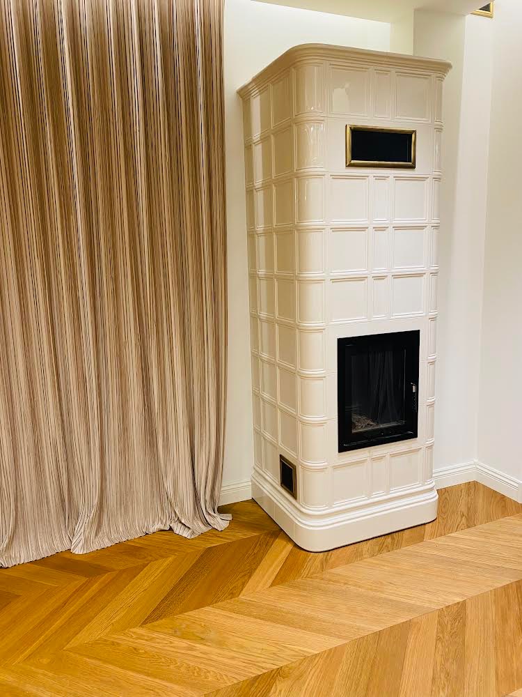 Biały, kaflowy piec z czarnym wkładem kominkowym, ustawiony na drewnianej podłodze ułożonej w jodełkę, obok beżowej, plisowanej zasłony.