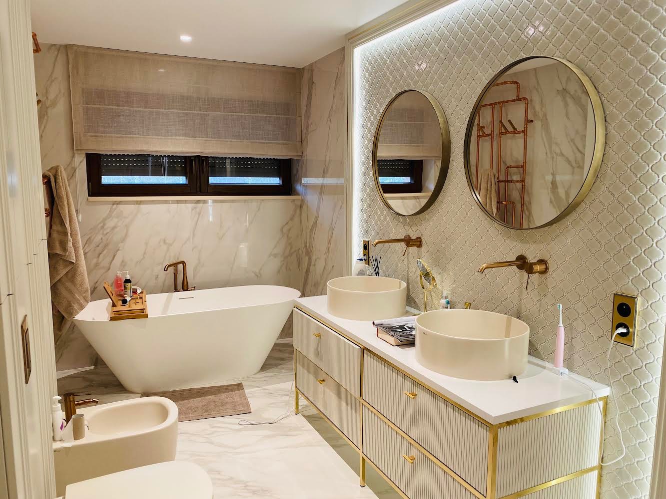 Elegancka łazienka z marmurowymi ścianami, owalną wanną wolnostojącą, dwiema okrągłymi umywalkami nablatowymi na białej szafce z dekoracyjnymi złotymi detalami, okrągłymi lustrami i mozaiką...