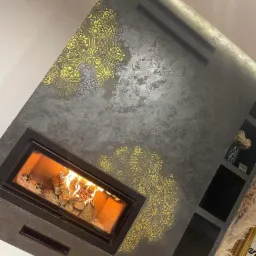 Szara ściana z dekoracyjnym tynkiem i złotymi mandalami, z wbudowanym kominkiem z palącym się drewnem oraz czarnymi półkami w ścianie.