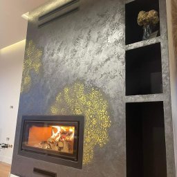 Beton.art - Ściana z dekoracyjnym tynkiem w odcieniach szarości, ozdobiona złotymi mandalami, z wbudowanym kominkiem z palącym się drewnem i wnękami. Na półce wazon z suszonymi kwiatami.