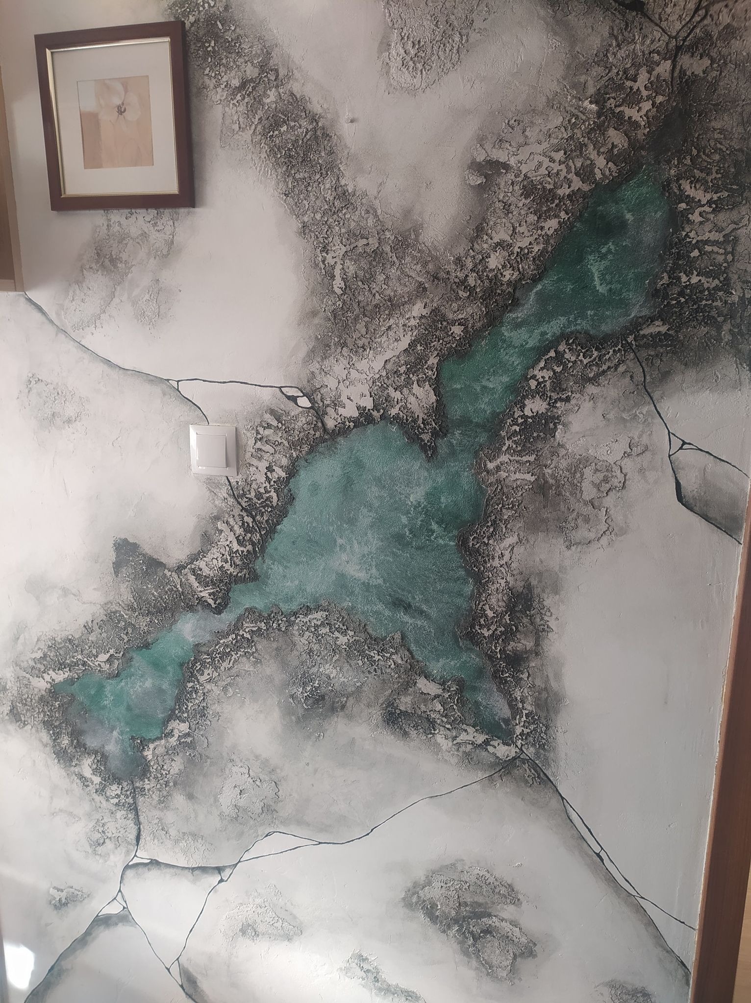 Dekoracyjna ściana z efektem abstrakcyjnego krajobrazu: turkusowe plamy przypominające jeziora otoczone szarymi, teksturowanymi obszarami. Na ścianie wisi obrazek w brązowej ramce, obok widoczny...