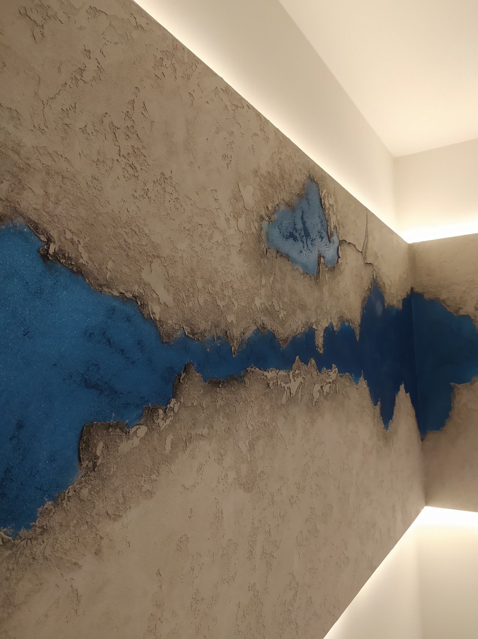 Ściana z dekoracyjną, teksturowaną farbą w odcieniach beżu i błękitu, przypominająca abstrakcyjny krajobraz z rzeką, oświetlona subtelnym światłem LED z sufitu.