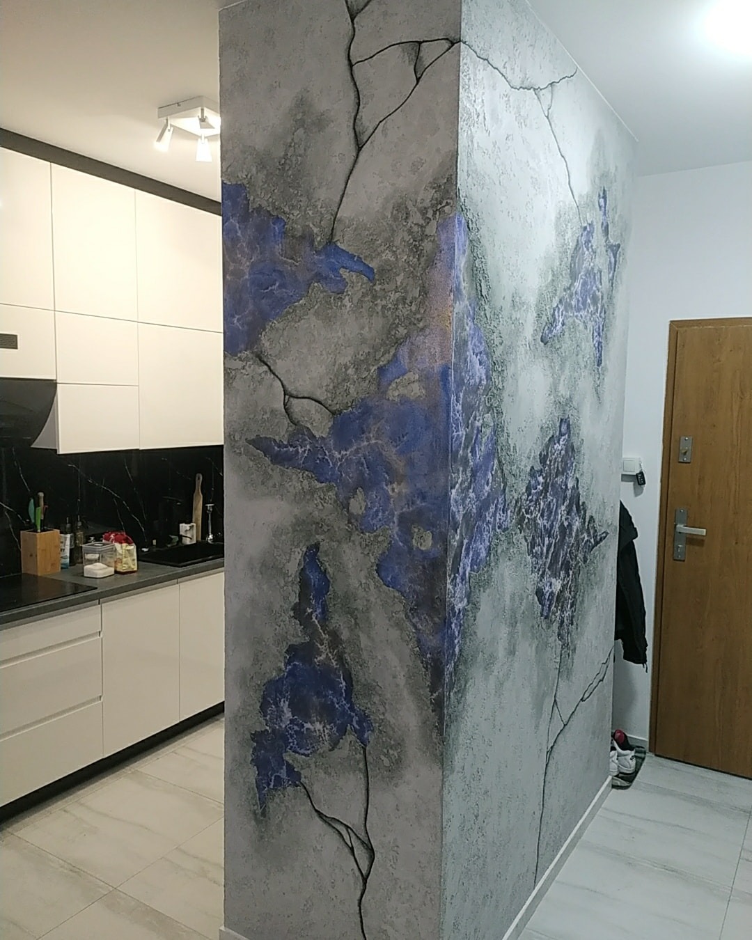Dekoracyjna ściana działowa w mieszkaniu, imitująca spękany kamień z niebieskimi akcentami, widok na kuchnię z białymi szafkami i czarnym blatem.