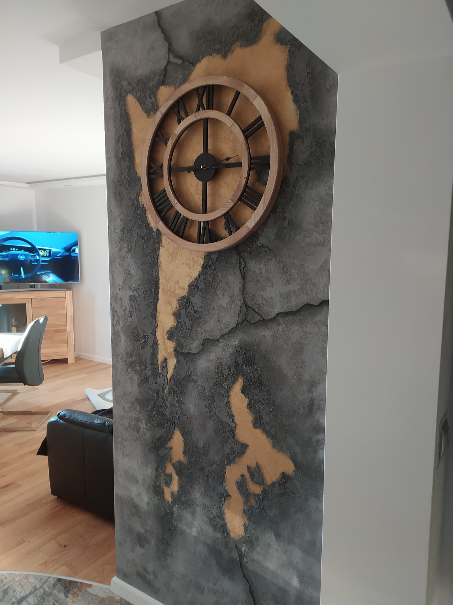 Dekoracyjna ściana z efektem spękanego tynku w odcieniach szarości i złota, z dużym drewnianym zegarem z rzymskimi cyframi, widok na salon z telewizorem w tle.