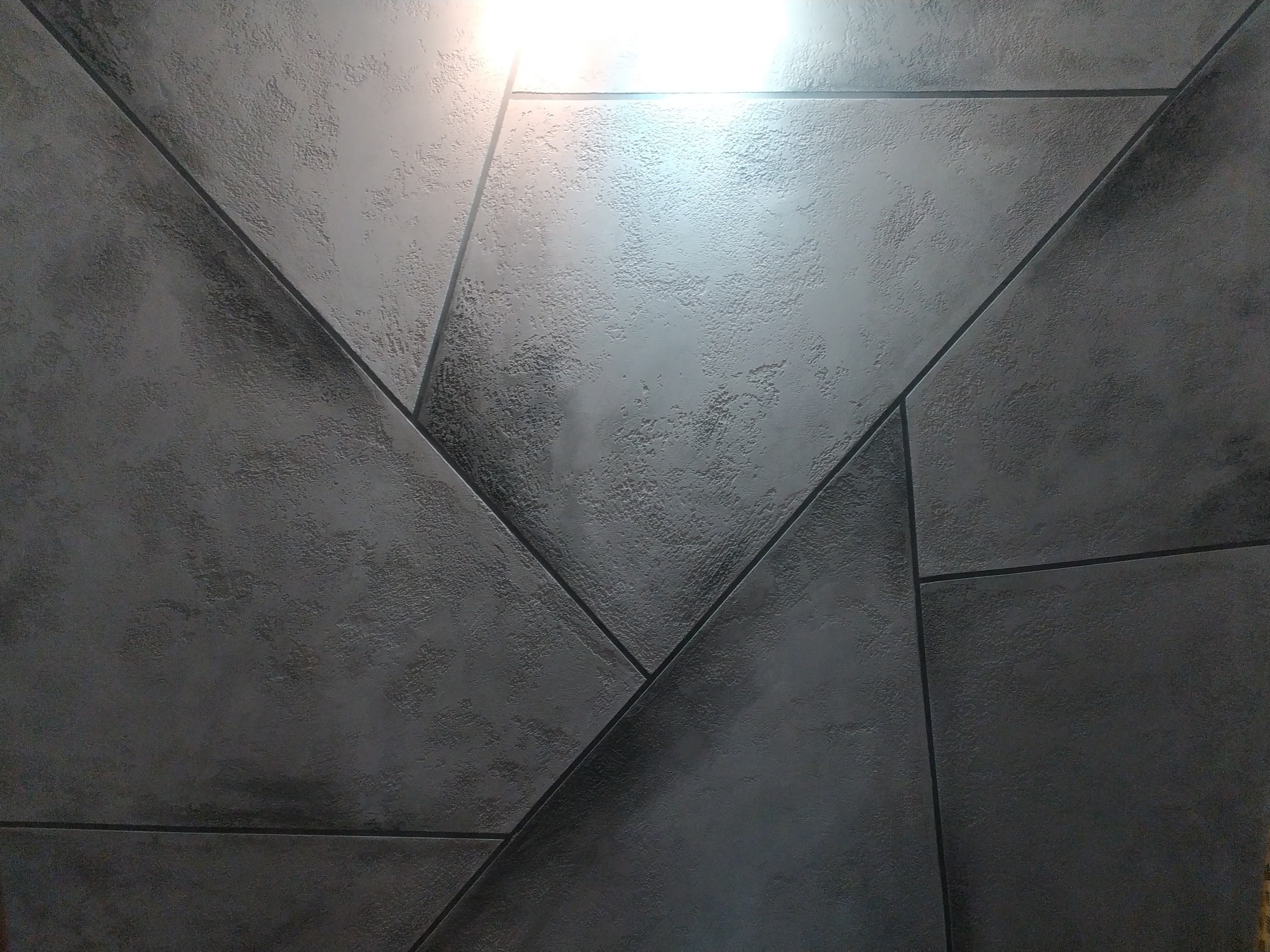 Dekoracyjna ściana w odcieniach szarości z geometrycznym wzorem, imitująca betonową strukturę, oświetlona od góry.