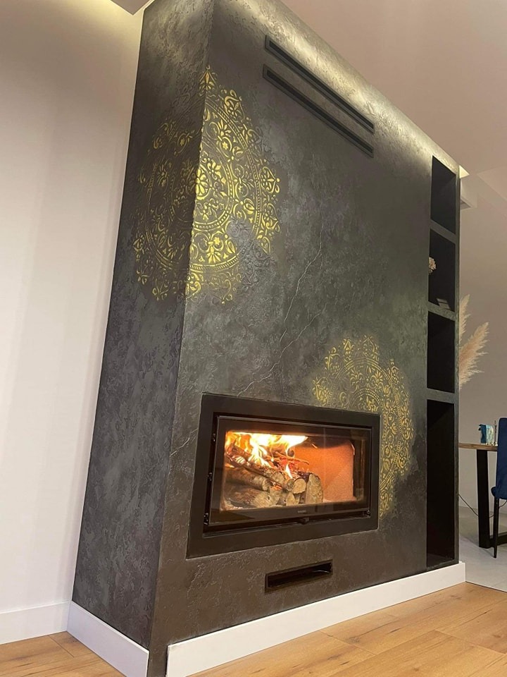 Nowoczesny kominek z czarną obudową, zdobiony złotymi, orientalnymi wzorami, z palącym się drewnem wewnątrz i półkami po prawej stronie.