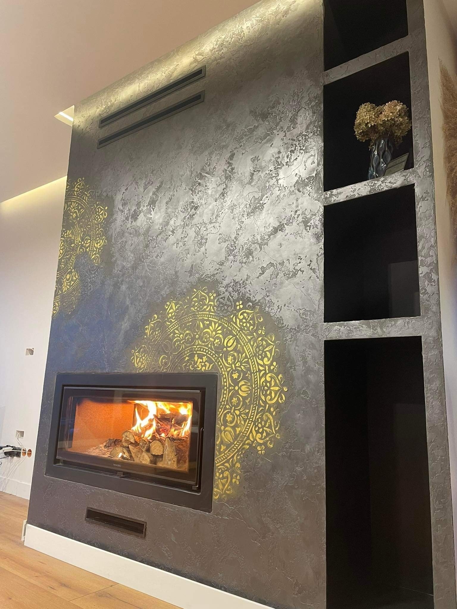 Ściana z dekoracyjnym tynkiem w odcieniach szarości, ozdobiona złotymi mandalami, z wbudowanym kominkiem z palącym się drewnem i wnękami. Na półce wazon z suszonymi kwiatami.