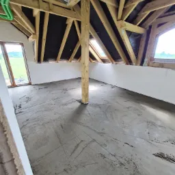 Poddasze z drewnianą konstrukcją dachu i wylaną, surową posadzką; widoczne okna dachowe i pionowy drewniany słup podtrzymujący konstrukcję.