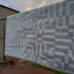 Szary dywan z geometrycznym wzorem, suszony na zewnątrz na metalowym stojaku przy szarej ścianie budynku i brązowym pojemniku na śmieci, pochmurne niebo w tle.