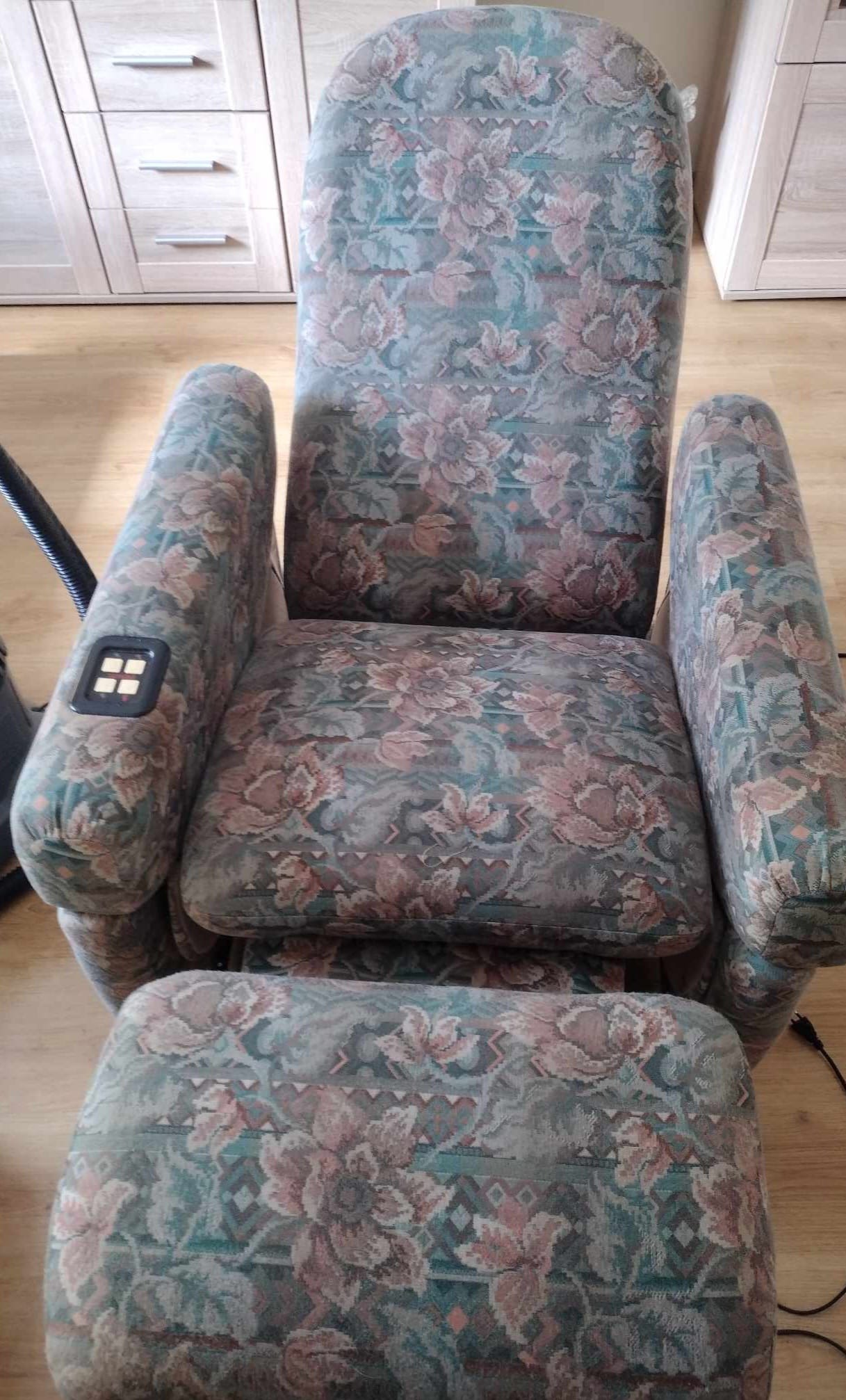 Tapicerowany fotel rozkładany z podnóżkiem w kwiatowy wzór, widoczne panele sterowania na podłokietniku, na drewnianej podłodze, z elementami odkurzacza w tle.