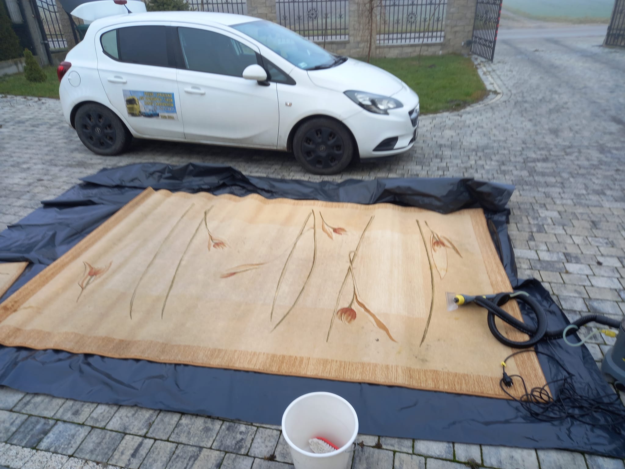 Beżowy dywan z kwiatowym wzorem rozłożony na folii, obok biały samochód i sprzęt do czyszczenia, na zewnątrz, kostka brukowa.