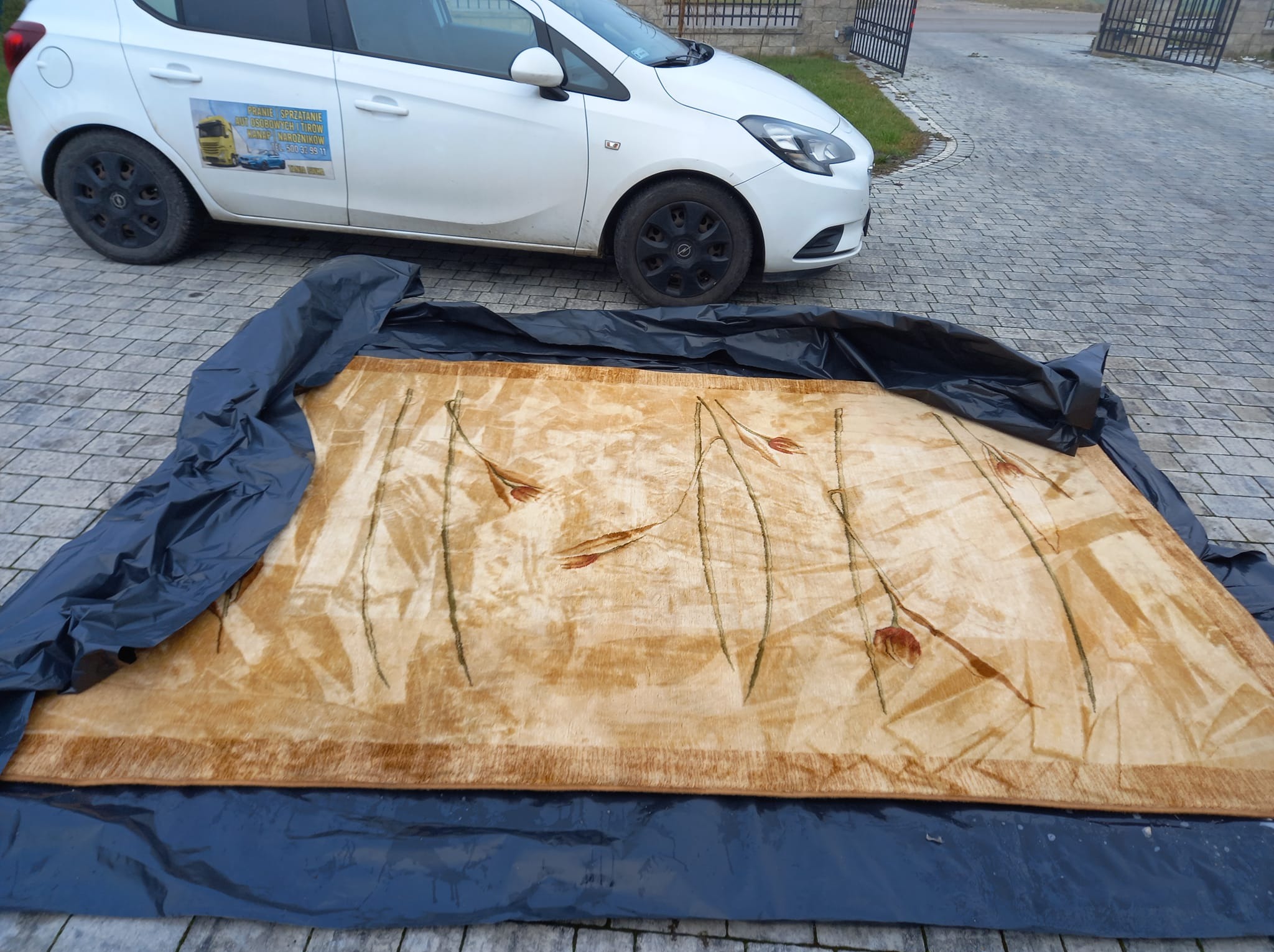 Beżowy dywan z motywem kwiatowym, rozłożony na czarnej folii ochronnej na zewnątrz, obok zaparkowany biały samochód z reklamą usług prania.