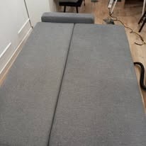 Rozłożona, szara kanapa z widocznymi śladami po praniu na jasnej podłodze. Fragment drabiny i krzesła w tle. Widok z góry.