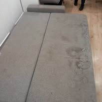 Szara, rozłożona kanapa z widocznymi, ciemniejszymi plamami na tapicerce. Widok z góry, fragment nóg osoby w tle.