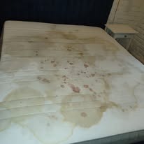 Zabrudzony materac z widocznymi plamami i zaciekami, wymagający profesjonalnego czyszczenia. Obok materaca stoi biała szafka nocna.