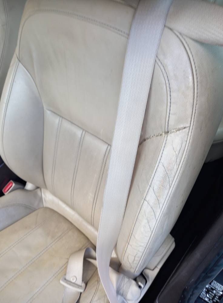 Beżowe skórzane siedzenie samochodowe z widocznymi zabrudzeniami i pęknięciami tapicerki, zapięty pas bezpieczeństwa. Perspektywa z góry, wnętrze pojazdu.