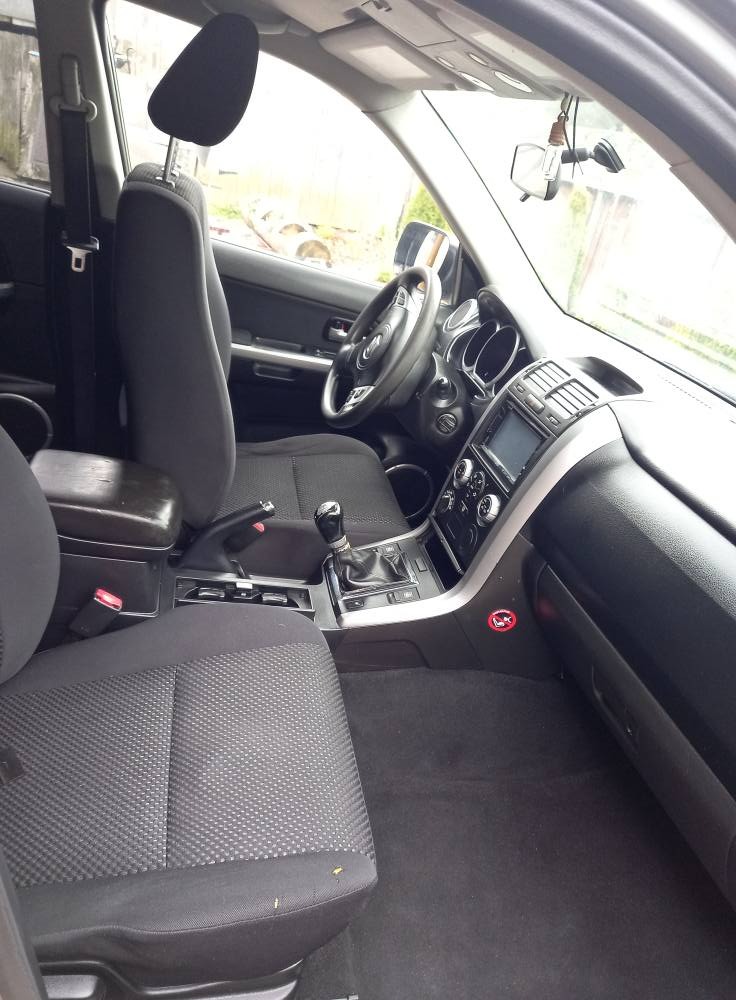 Wnętrze samochodu z widocznymi fotelami z ciemną tapicerką, deską rozdzielczą i konsolą środkową. Widoczny fragment kierownicy i przedniej szyby.