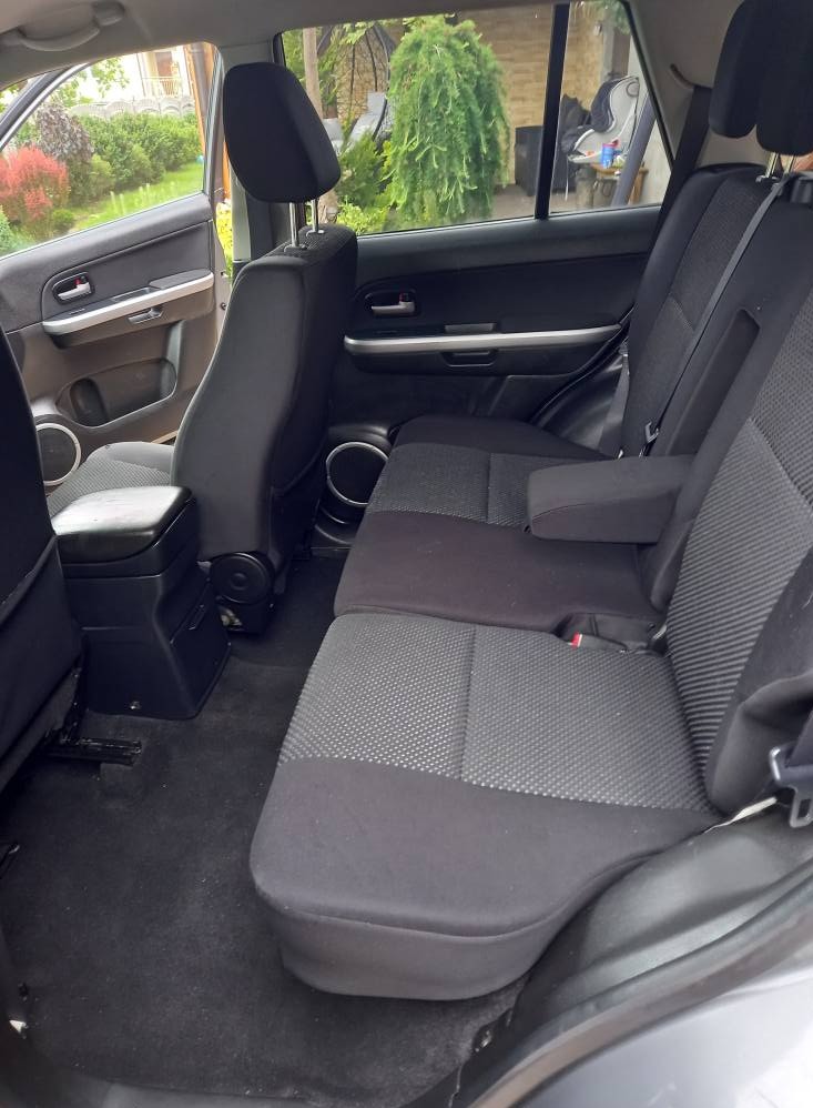 Wnętrze samochodu z czarną tapicerką, widok na tylną kanapę z podłokietnikiem i zagłówkami. Widoczny fragment drzwi i otoczenie za oknem.