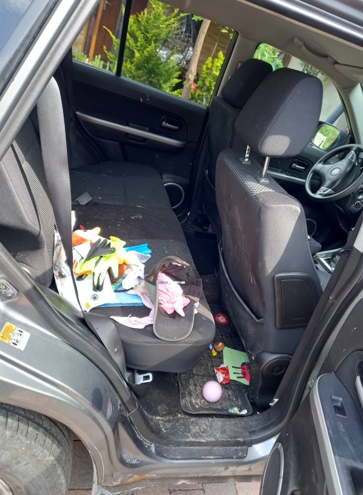 Wnętrze brudnego samochodu z otwartymi drzwiami. Na tylnych siedzeniach i podłodze widoczne śmieci, zabawki i inne przedmioty. Tapicerka wymaga gruntownego czyszczenia.