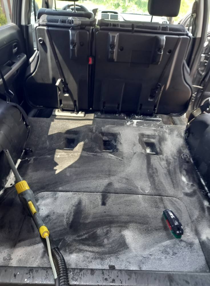 Czyszczenie tapicerki samochodowej z użyciem odkurzacza piorącego. Widoczne ślady piany i detergentu na dywanikach. Detailing wnętrza pojazdu.