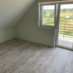 Poddasze po remoncie z ułożonymi panelami podłogowymi w odcieniach szarości, widok na zewnątrz przez przeszklone drzwi balkonowe z metalową barierką.