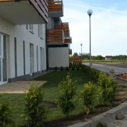 Nowo założony trawnik z niskimi tujami wzdłuż chodnika przed budynkiem apartamentowym z drewnianymi balkonami. W tle widoczne lampy uliczne i przygotowany teren pod parking.