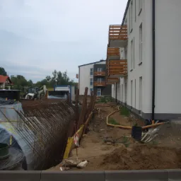 Wykonanie wykopu pod fundamenty budynku mieszkalnego, widoczne zbrojenia, szalunki oraz ciężki sprzęt budowlany w tle.