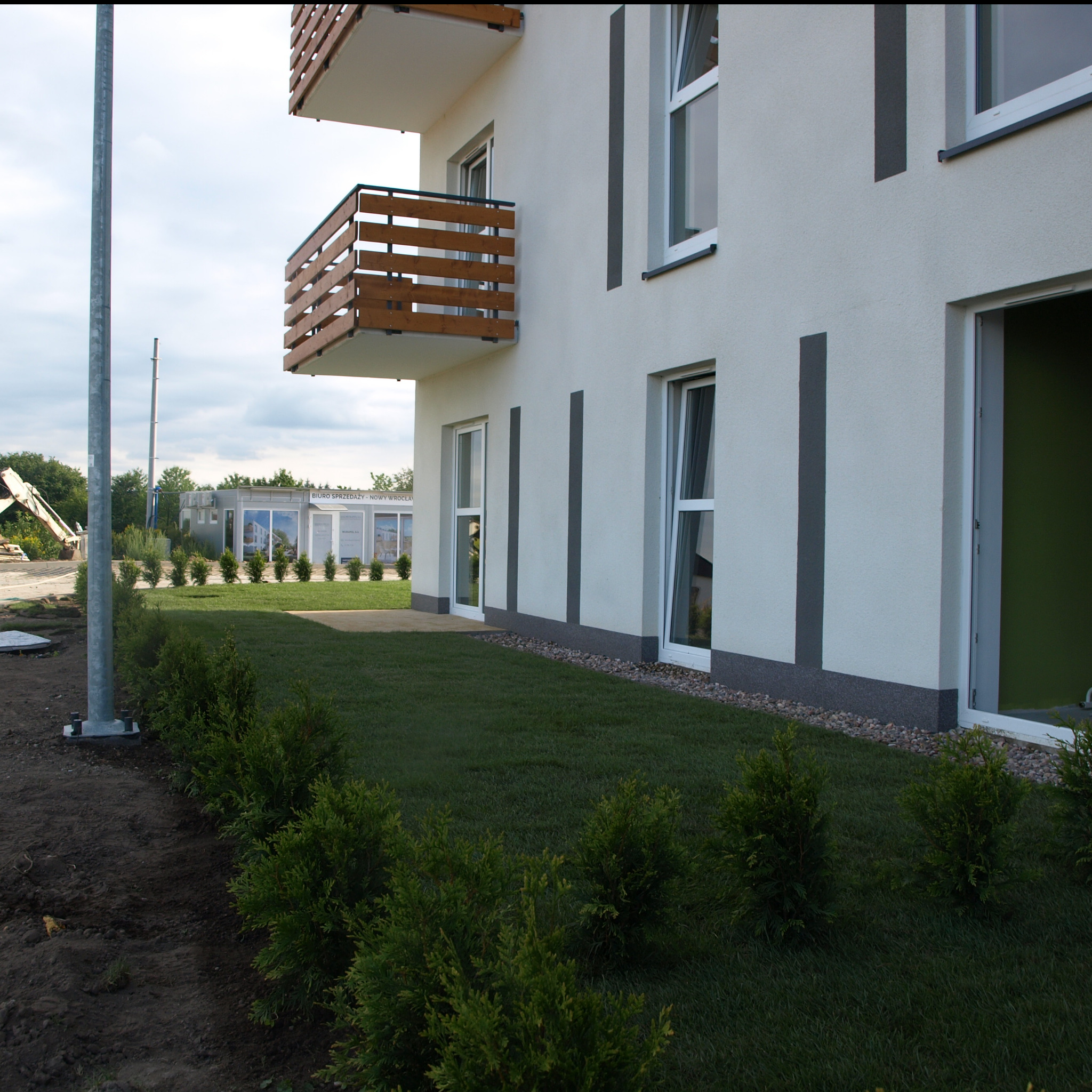 Nowo założony trawnik z rolki i zasadzone małe tuje wzdłuż ścieżki, przy białym budynku z drewnianymi balkonami.