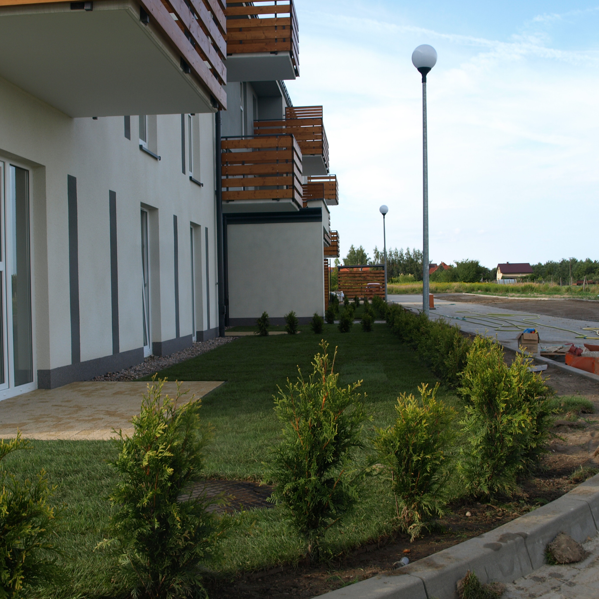 Nowo założony trawnik z niskimi tujami wzdłuż chodnika przed budynkiem apartamentowym z drewnianymi balkonami. W tle widoczne lampy uliczne i przygotowany teren pod parking.