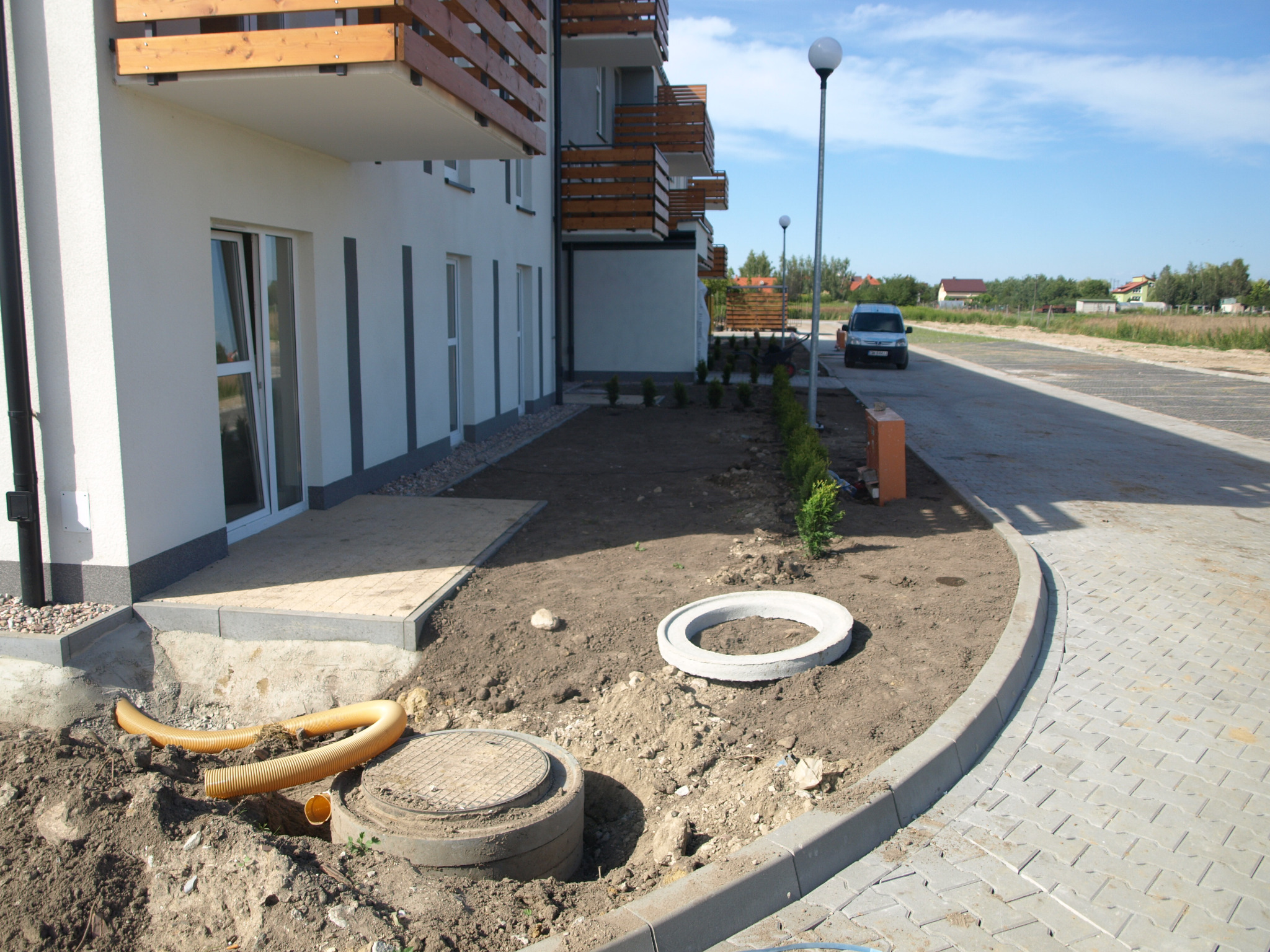 Teren przed nowym budynkiem z wybrukowanym chodnikiem, przygotowany grunt pod przyszły ogród z widocznymi elementami kanalizacji i nasadzeniami młodych krzewów.