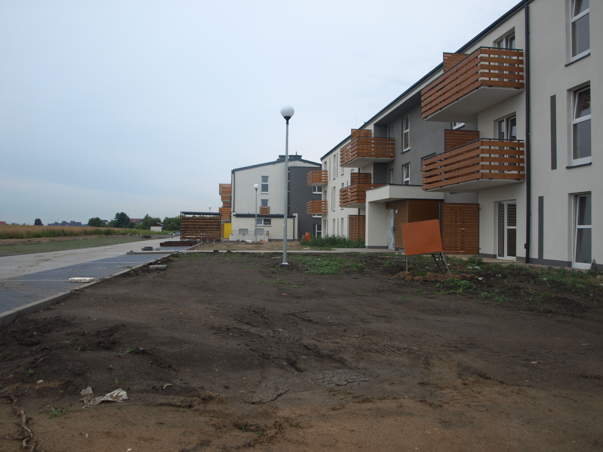 Przygotowanie terenu pod założenie ogrodu przed nowoczesnym budynkiem mieszkalnym z drewnianymi balkonami, widoczne ślady opon i lampa oświetleniowa.