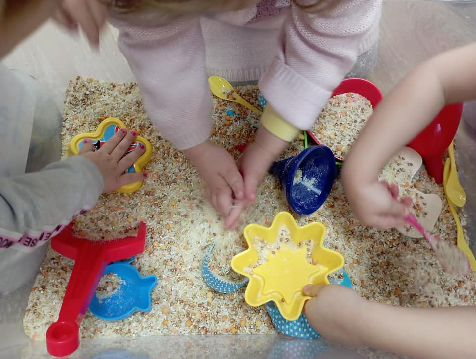 Dzieci bawią się w sensorycznym pudełku z ryżem i kolorowymi zabawkami, w tym łopatkami, foremkami w kształcie gwiazd i kubeczkami.