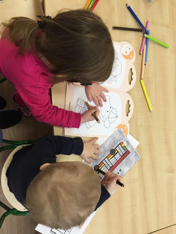 Dwoje dzieci, dziewczynka w różowej bluzce i chłopiec w granatowej, siedzą przy jasnym drewnianym stole i kolorują obrazki kredkami; dziewczynka koloruje słonia, a chłopiec autobus.