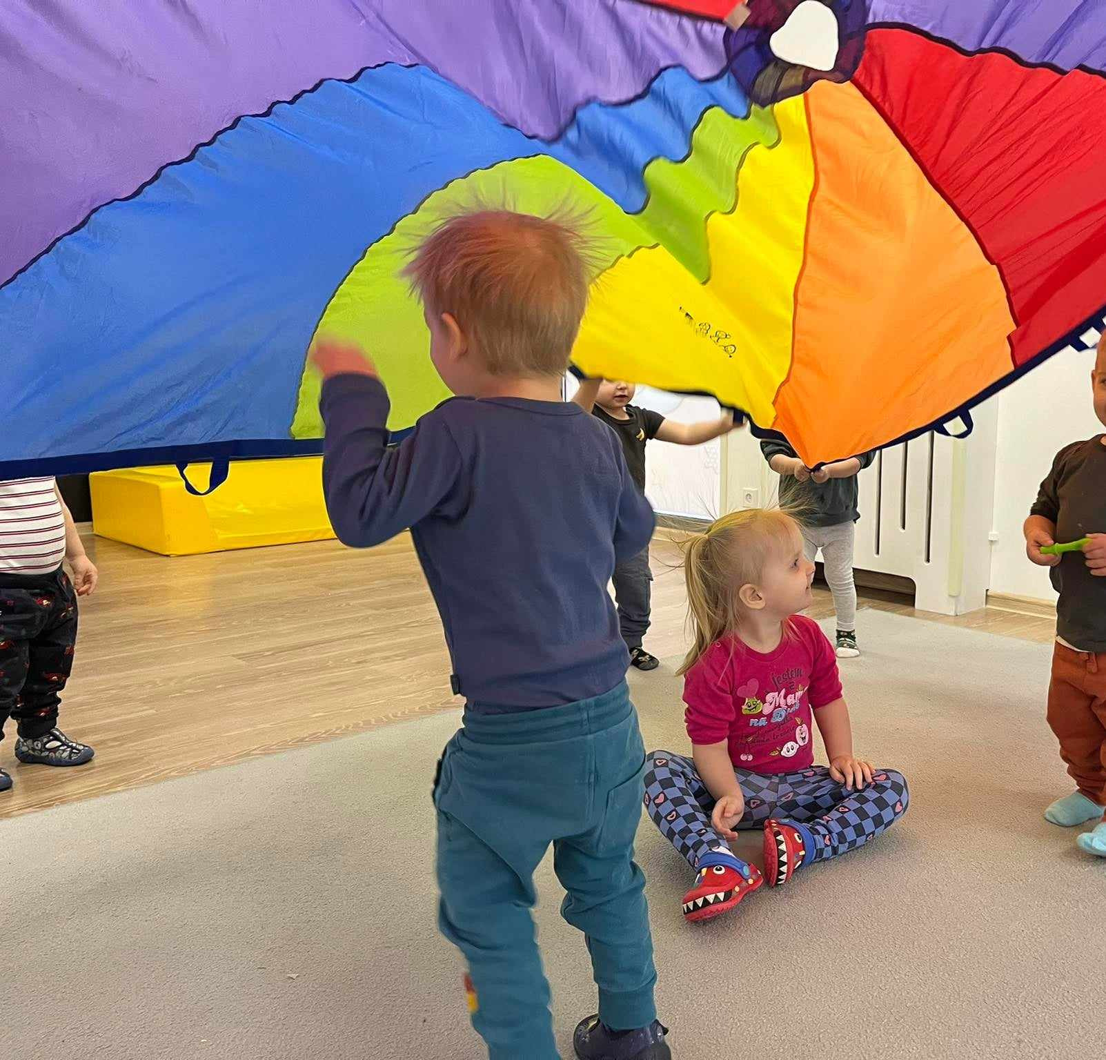 Dzieci bawią się pod kolorową chustą animacyjną w kształcie spadochronu, w tle widoczny żółty element wyposażenia sali zabaw.