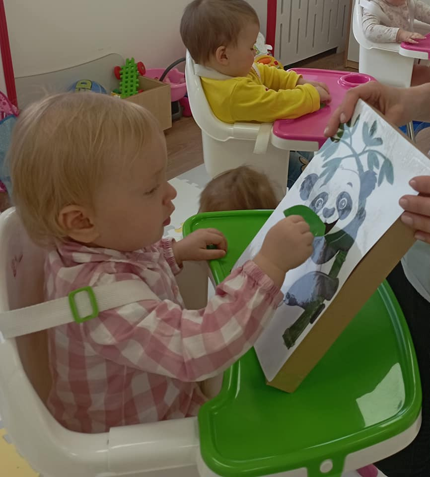 Malutka dziewczynka w kraciastej koszuli siedzi w krzesełku do karmienia, trzymając zielony element przy kartonowej ramce z rysunkiem pandy, w tle inne dzieci.