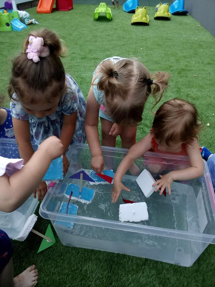 Trójka małych dzieci bawi się na zewnątrz w przezroczystym pojemniku z wodą i pływającymi, ręcznie robionymi łódkami z pianki i papieru, na tle zielonego trawnika z zabawkami.