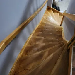 Drewniane schody kręcone podczas cyklinowania, widoczne ślady pyłu po obr&oacute;bce, jasna barierka, szara ściana w tle.