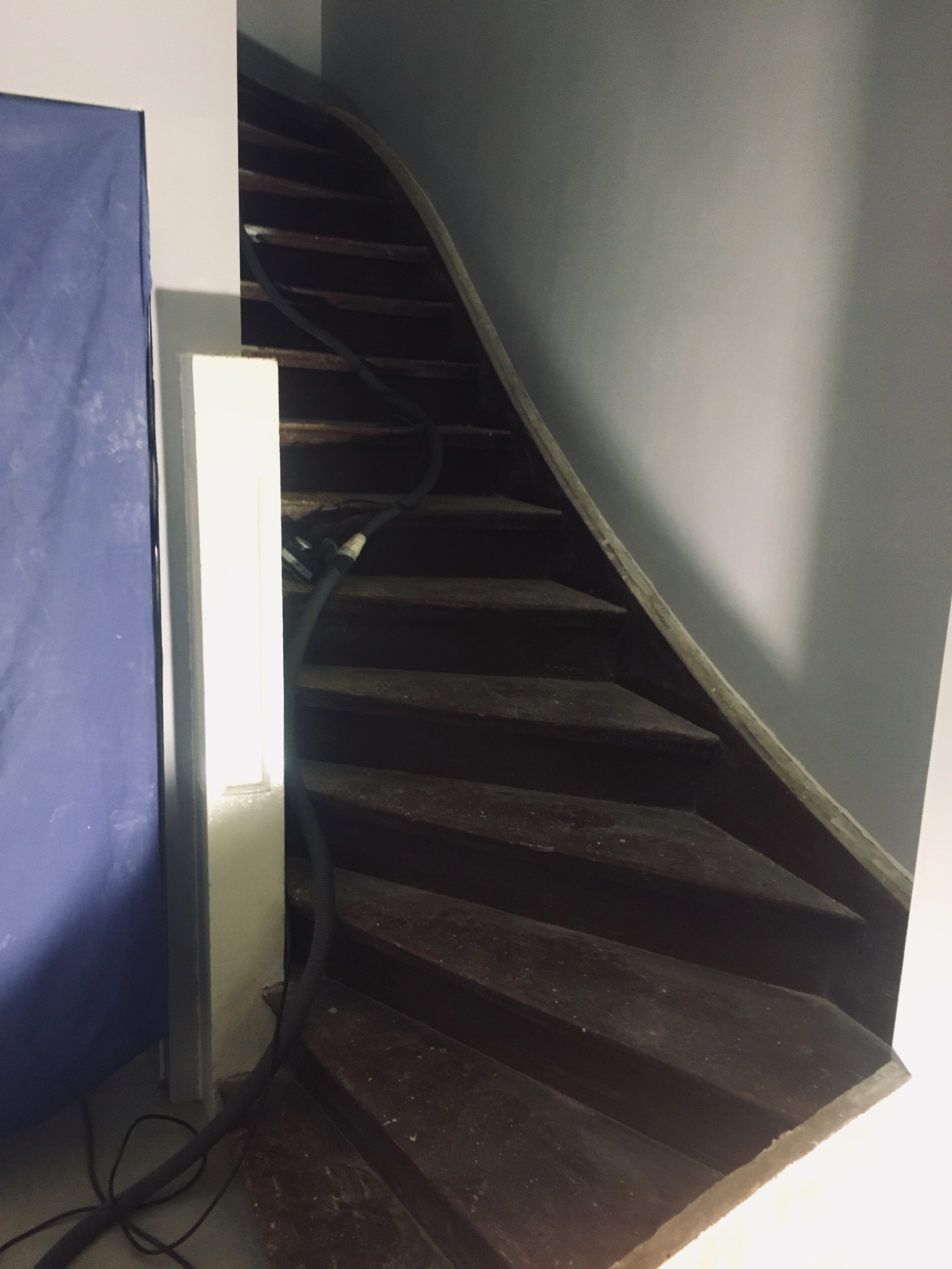 Ciemne drewniane schody kręcone w trakcie cyklinowania, widoczny wąż od odkurzacza, szare ściany i niebieska folia ochronna.