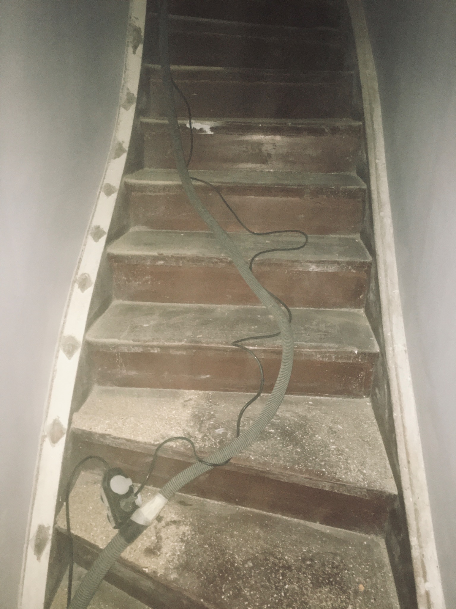 Drewniane schody w trakcie renowacji, pokryte pyłem, z widocznym wężem odkurzacza przemysłowego, przygotowane do cyklinowania.