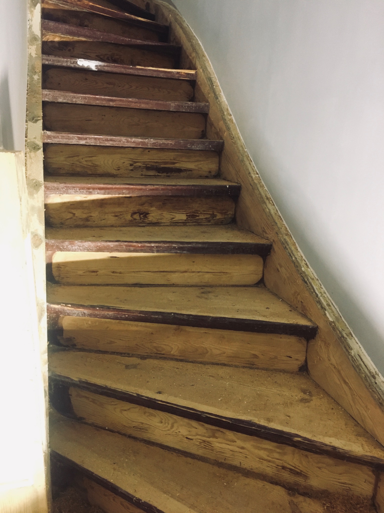 Drewniane schody w trakcie renowacji, z widocznymi śladami starej farby i pyłu po szlifowaniu, oświetlone z góry, ukazujące teksturę drewna i niedoskonałości.