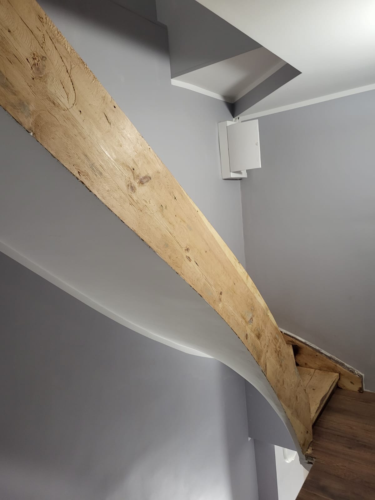 Drewniana belka stropowa, prawdopodobnie po cyklinowaniu, w nowoczesnym, szarym wnętrzu. Widoczna struktura drewna i fragment parkietu na schodach.