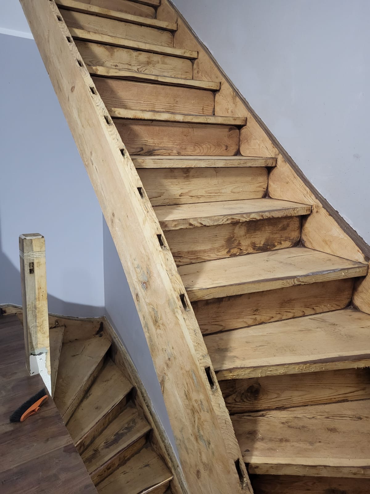 Drewniane schody z surowego drewna, w trakcie renowacji, widoczne ślady po cyklinowaniu, z boku leży szczotka do zamiatania.