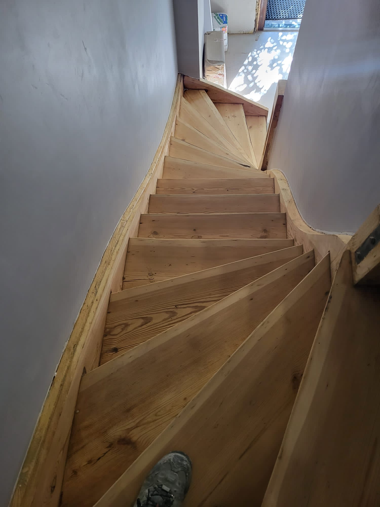 Wewnętrzne drewniane schody kręcone, surowe, z widoczną strukturą drewna, świeżo po montażu, z szarą ścianą po bokach i butem roboczym w dolnym rogu.
